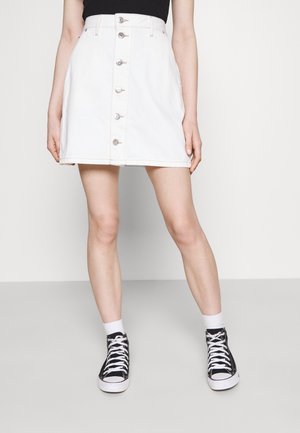 Jupe en denim blanc avec boutons sur le devant, taille cintrée et ourlet droit. Associée à des baskets montantes noires et des chaussettes blanches.