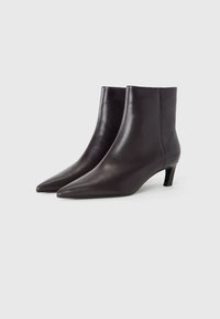 Une paire de bottines en cuir marron foncé avec des pointes aiguës et des talons courts sur un fond blanc uni.