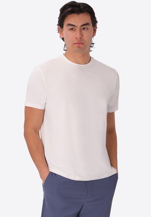 Uomo con capelli scuri che indossa una semplice maglietta bianca a maniche corte e pantaloni blu con le mani nelle tasche, che guarda leggermente di lato.