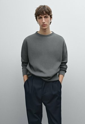 Pull gris à côtes avec un col rond et des manches longues, associé à un pantalon plissé bleu marine foncé. Le modèle se tient devant un fond uni.