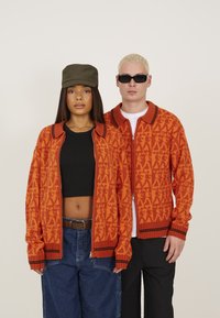 Deux mannequins portant des cardigans en maille orange assortis avec un design à motifs, des accents noirs et des tenues contrastées. L'un porte un haut noir et une casquette.