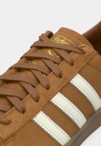 Brūni zamšaini sporta apavi ar bēšiem ādas svītrām, plakaniem brūniem auklām un zelta logo uz mēles. Tekstūrēta virsma un noapaļota forma.