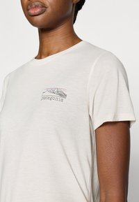 Μπεζ βαμβακερό t-shirt με υφασμάτινη υφή, στρογγυλή λαιμόκοψη και σχέδιο βουνού με τη λέξη "patagonia" στο ύψος του στήθους.