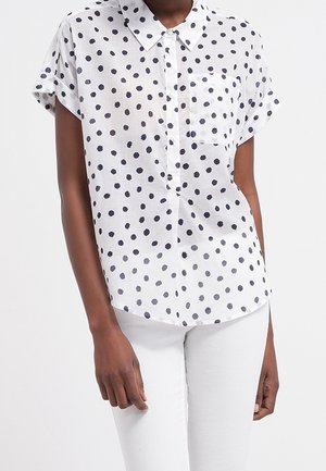 Woman wearing a white short-sleeve button-up shirt with black irregular polka dots and white pants, standing against a plain background.