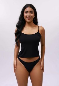 Νεαρή γυναίκα με μακριά σκούρα μαλλιά, φορώντας μαύρη καμισόλα και ταιριαστά μπικίνι, στέκεται και χαμογελάει μπροστά σε απλό φωτεινό φόντο.