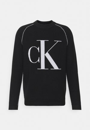Černý svetr s velkým bílým logem "CK" na přední straně. Kulatý výstřih, dlouhé rukávy a kontrastní švy na ramenou.