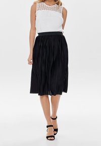 Gonna nera plissettata con vita aderente, abbinata a un top senza maniche bianco con dettaglio in pizzo sul collo. Sandali neri con tacco.