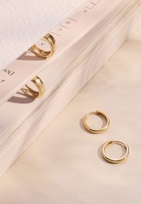 Boucles d'oreilles en or avec une surface lisse et brillante. Deux boucles d'oreilles sont exposées ; l'une est partiellement cachée derrière une serviette blanche texturée et un livre.