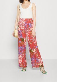 Czerwone szerokie spodnie w kwiaty z kolorowymi niebieskimi i białymi kwiatami. Połączone z białym prążkowanym topem na ramiączkach i zielonymi sandałami na szpilce. Beżowa torebka.