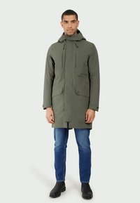 Zaļa virsjakas ar kapuci, priekšējais rāvējslēdzis un divas pogu kabatas. Ietver taisnu griezumu, mīkstu tekstūru un minimālistisku dizainu.