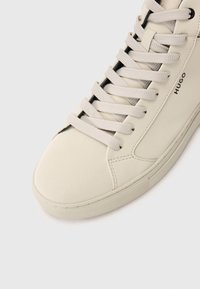 Beige sneaker met een ronde neus, gestructureerde veters, gestikte details en "HUGO" branding aan de zijkant, tegen een eenvoudige witte achtergrond.