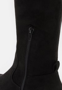 Primer plano de una bota de ante negro con una cremallera lateral vertical y un tirador en la parte trasera superior.