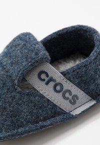 Chausson Crocs bleu en peluche avec sangle réglable portant l'inscription "crocs" sur un fond blanc, montrant un intérieur doux.