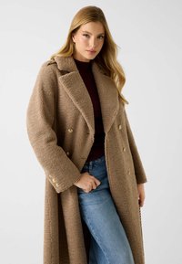 Brauner, strukturierter, langer Mantel mit zweireihigem Design und goldenen Knöpfen. Getragen über einem dunklen Oberteil, kombiniert mit hellblauen Jeans.