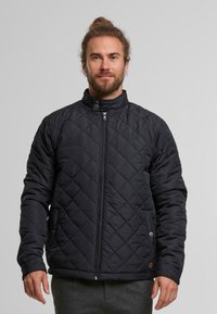 Czarna pikowana kurtka z wysokim kołnierzem, zamkiem błyskawicznym z przodu oraz kieszeniami po bokach. Gładka tekstura z wzorem w diamenty.