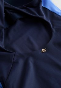 Tejido atlético azul marino con escote de malla, que presenta un acento en oro del logo. Textura suave, diseñado para el rendimiento y la comodidad.