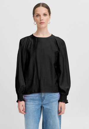 Blusa nera a maniche lunghe con scollo rotondo, spalle a palloncino e polsini arricciati. Tessuto liscio e vestibilità comoda. Indossata con jeans blu.