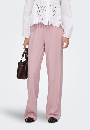 Femme portant un pantalon rose ample, un chemisier blanc à œillets, tenant un sac à main marron foncé texturé et des chaussures noires sur fond uni.