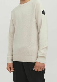 Beige strukturerad stickad tröja med rund hals och svart cirkulär lapp på den övre ärmen, bärs med svarta byxor med vit text.