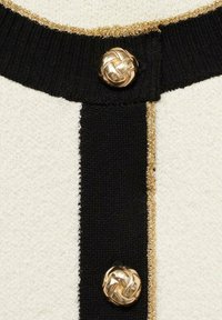 Tissu en tricot noir et blanc avec des bordures dorées ; présente de grands boutons en métal doré tressés au niveau du col.