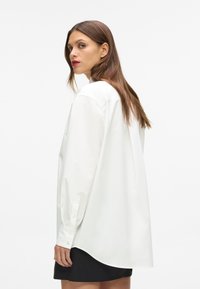 Biała, długosleeve'owa koszula z gładkiego materiału, charakteryzująca się luźnym krojem, zapięciami na guziki przy mankietach oraz zaokrąglonym dołem. Noszona z czarną spódnicą.