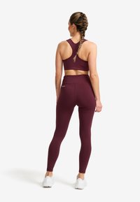 Kvinna med flätat hår som bär en vinröd sport-bh och leggings, stående med ryggen mot kameran mot vit bakgrund.