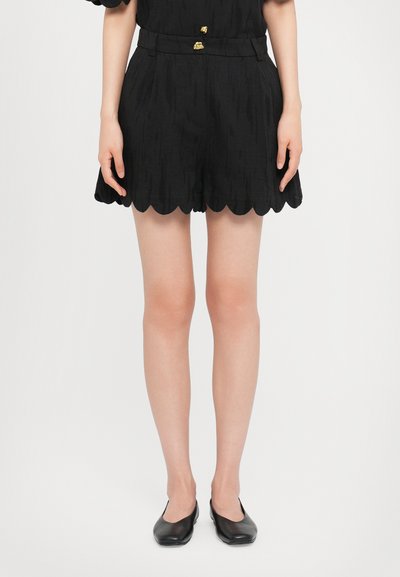 Sorte shorts med bølget kant i tekstureret stof, med en tætsiddende talje med guldhardware. Sammen med sorte flade sko.