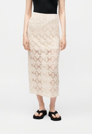 Femme portant une jupe midi beige en dentelle sur doublure couleur nude, un top noir sans manches, et des sandales compensées noires à lanières entre les orteils, debout sur fond blanc.