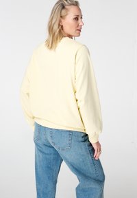 Gele trui met geribbelde manchetten, ontspannen pasvorm en ronde hals. Gecombineerd met lichtblauwe denim jeans. Achtergrond is effen wit.