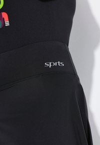 Zwarte sportkleding met een gladde textuur, een nauwsluitend ontwerp en een wit logo met de tekst "sports" op de tailleband. Levendige grafische accenten aanwezig.