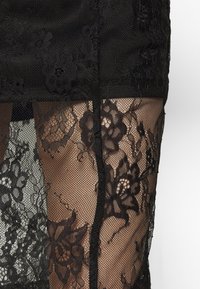 Svart genomskinlig spets med blommönster som ligger över huden, vilket visar en närbild av plaggens intrikata textur och design.