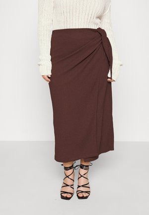 Kvinde iført en brun midi-nederdel med wrap-stil og sidesløjfe, hvid striktrøje og sorte sandaler med hæl og stropper.