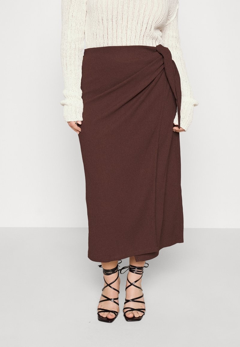 Femme portant une jupe midi portefeuille marron avec un nœud sur le côté, un pull en maille blanc et des sandales à talons hauts noires à lanières.