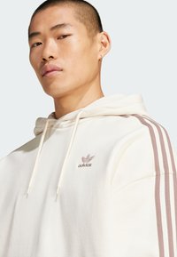 Krems krāsas kapuce ar aukliņu un trim rozā svītrām uz katras piedurknes. Uz krūtīm iešūts Adidas logs. Maiga tekstūra.