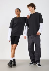 Dwa modele noszą kontrastujące stylizacje: czarny prążkowany top z białymi rękawami, czarne szorty i szare spodnie. Zauważalne buty i sneakersy dopełniają ich wygląd.