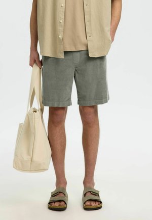 Mann trägt beigefarbenes kurzärmeliges Hemd, hellbraunes T-Shirt, graue Cordshorts, olivfarbene Sandalen und hält eine große cremefarbene Canvas-Tragetasche.