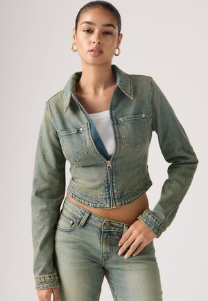 Jeune femme portant une veste en jean courte bleu clair sur un haut blanc, un jean taille haute assorti, et des boucles d'oreilles créoles dorées, posant devant un fond uni.