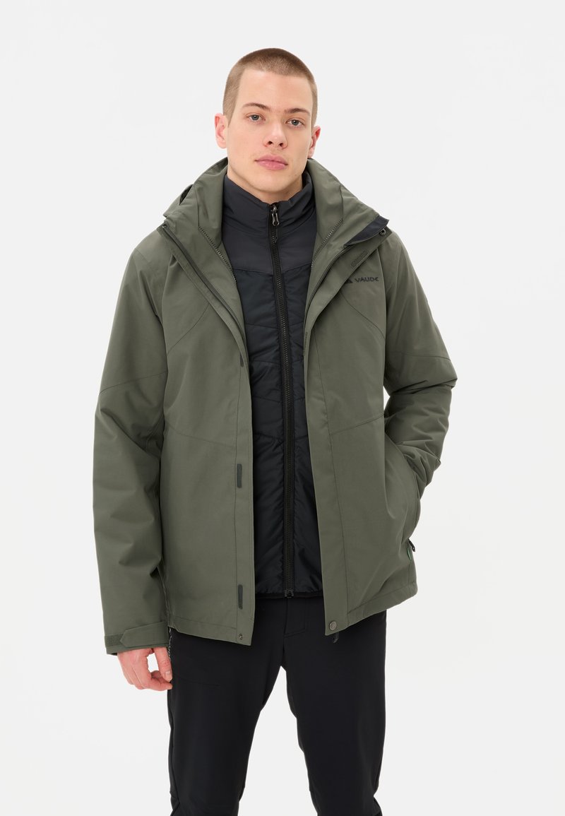 Homme portant des vestes d'extérieur superposées, une veste extérieure verte olive avec capuche et une veste intérieure rembourrée noire, se tenant contre un fond blanc.
