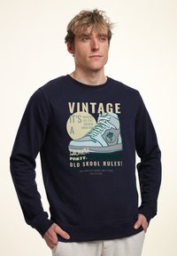 Felpa navy con grafica di una sneaker, dettagli testuali in crema e verde, polsini e orlo a coste, realizzata in un materiale morbido.