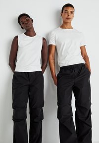 Δύο άτομα στέκονται δίπλα-δίπλα. Φορούν λευκά βαμβακερά πάνω μέρη και μαύρο νάιλον παντελόνια με χαλαρή εφαρμογή και ελαστική μέση.