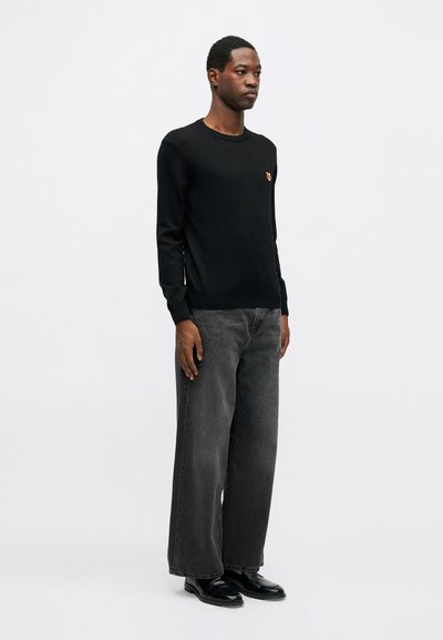Mann i svart genser med liten rev-emblem, vide grå jeans og svarte lærsko som står mot hvit bakgrunn.