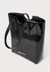 Saco tote preto feito de couro sintético brilhante e envernizado, com duas alças longas, apresentando um design minimalista e um detalhe em logo prateado.