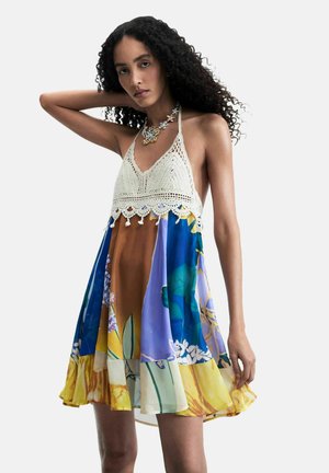 Vrouw met krullend haar die een mouwloze halterjurk draagt met een witte gehaakte top en een meerkleurige bloemenrok, poserend met één arm omhoog.