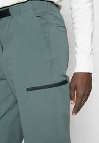 Höger hand bredvid grå byxor med svart dragkedjeficka, bärs med vit långärmad skjorta synlig vid handleden