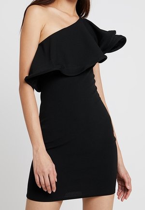 Vrouw draagt een mouwloze zwarte mini-jurk met een grote volantdetail over één schouder en over de borst.