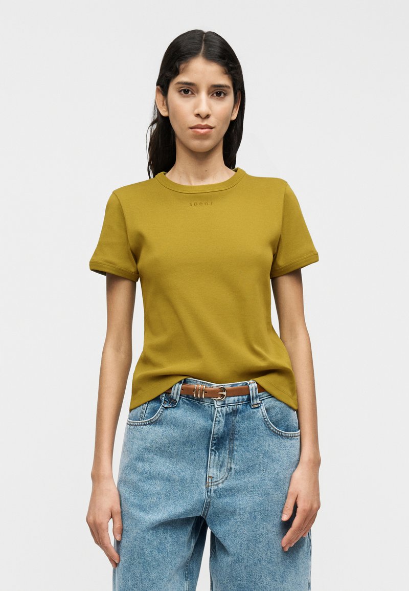 Λαδί πράσινο κοντομάνικο μπλουζάκι με ριμπ γιακά και μικρή λεπτομέρεια λογότυπου, συνδυασμένο με ψηλόμεσο ανοιχτό μπλε τζιν παντελόνι.