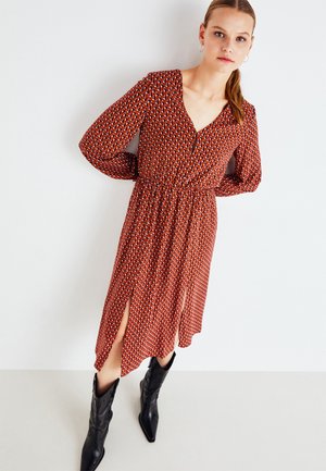 Κόκκινο φόρεμα με σχέδιο, μακριά μανίκια, V-λαιμόκοψη και συγκεντρωμένη μέση. Διαθέτει πλαϊνές σχισμές και είναι συνδυασμένο με μαύρες μυτερές μπότες.