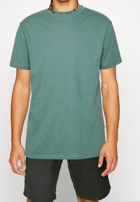 T-shirt en coton vert pâle, col rond, manches courtes, coupe décontractée, texture lisse avec étiquette sur la couture latérale visible.