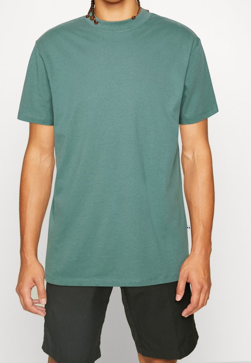 T-shirt en coton vert pâle, col rond, manches courtes, coupe décontractée, texture lisse avec étiquette sur la couture latérale visible.