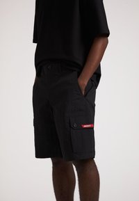 Schwarze Cargo-Shorts aus strukturiertem Stoff, mit zwei Seitentaschen, einer mit einem Knopf verschlossenen Tasche mit einem roten "JACKIT"-Label und einem Bund mit Knöpfen.